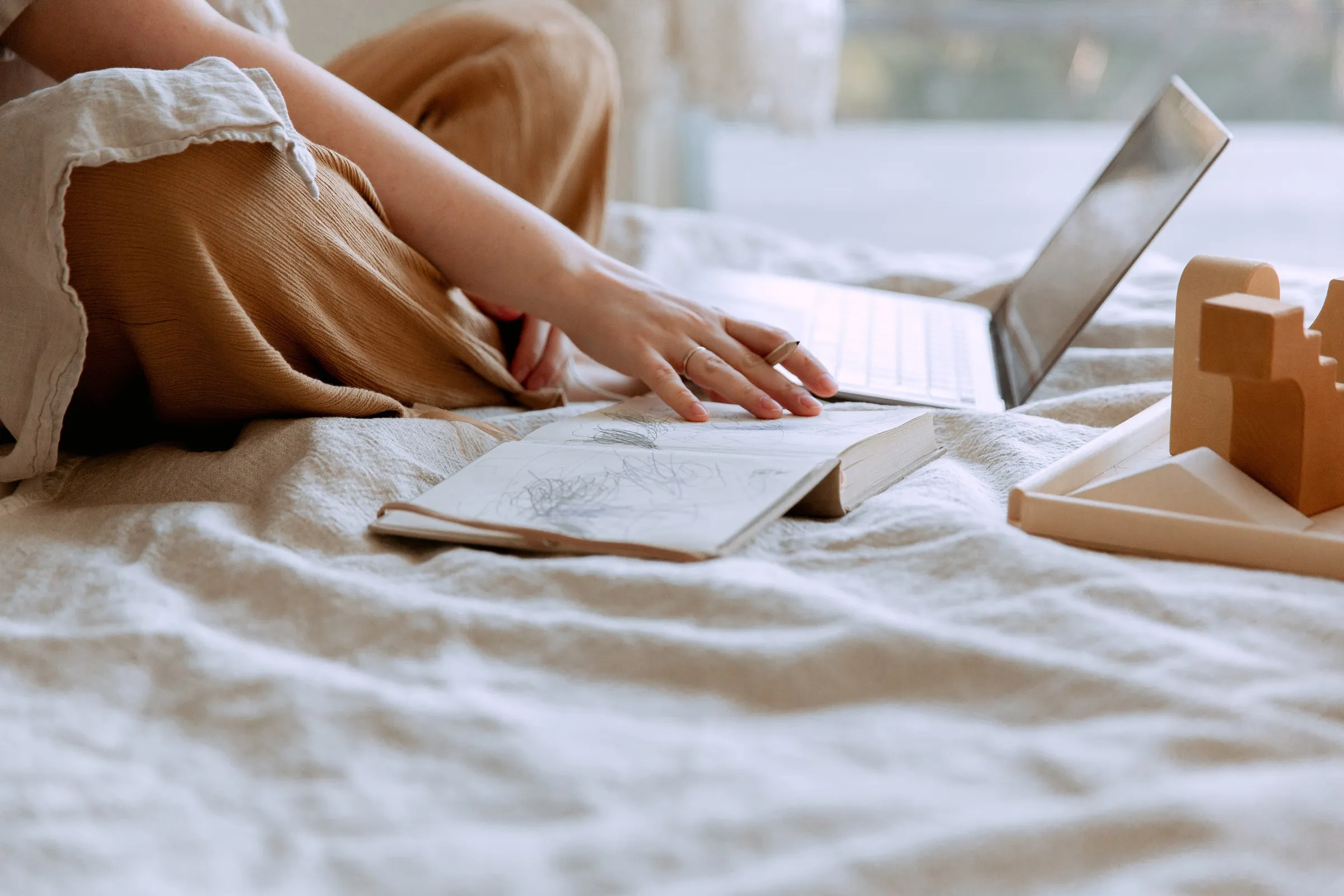 Frau sitzt mit einem aufgeschlagenen Skizzenbuch und einem Laptop auf einem Bett und blättert in ihren Notizen